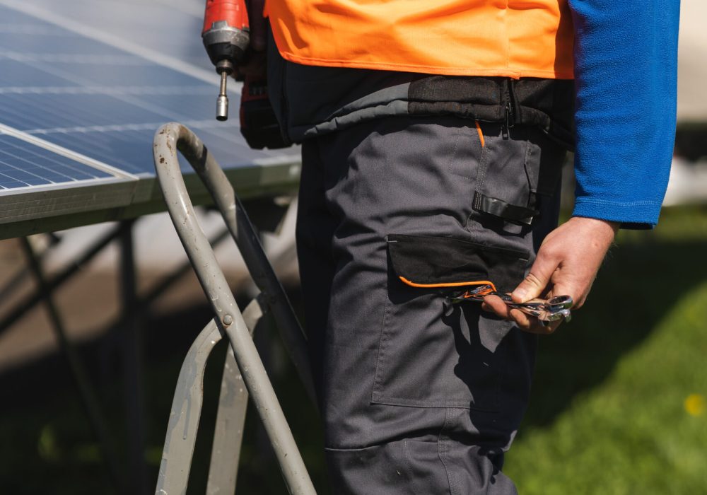 Male engineer who installs and maintains solar panels stands on a stepladder and takes out a wrench from his pocket